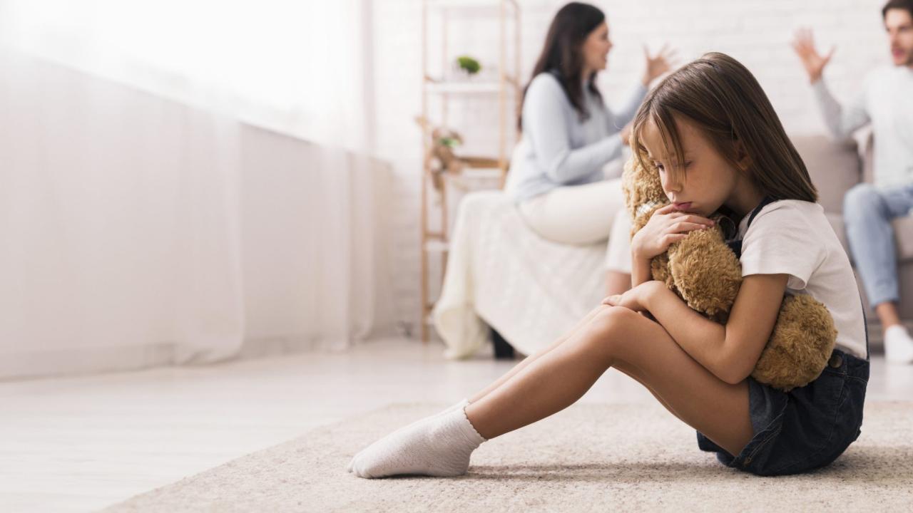 nena abraçada a un peluix amb pares discutint de fons
